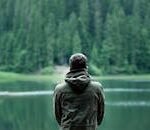 A person in a green jacket stands by a serene forest lake, capturing calming nature.