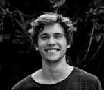 A black and white portrait of a young man smiling outdoors with natural background.